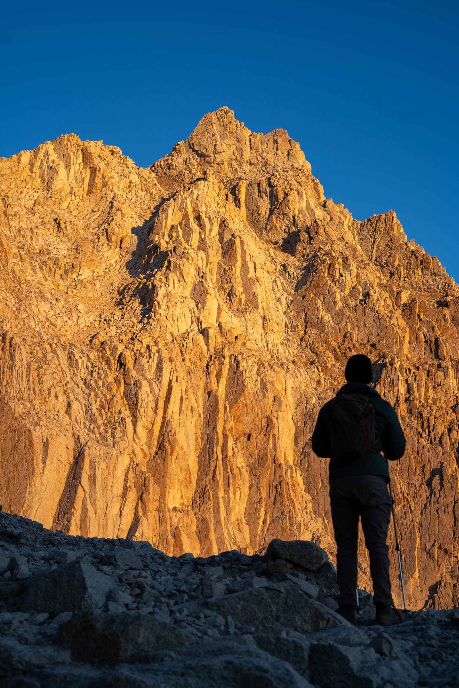 A hiker standing in silhouette at the base of a golden sunlit granite face